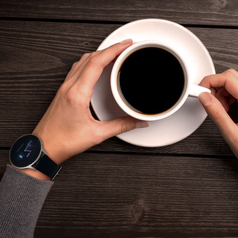Person holding a white cup of coffee on a wooden table with a smartwatch on the wrist.