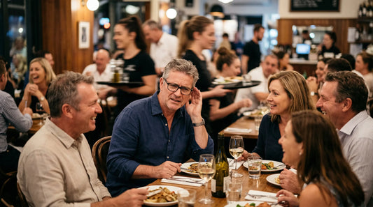 Gemini said A busy restaurant scene where five middle-aged adults are dining together, smiling and talking. The man in the center wears glasses and has his hand raised to his ear.
