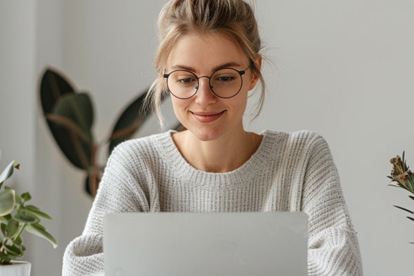 Woman wearing glasses and a sweater, using a laptop indoors with plants in the background